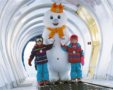Zwei Kinder stehen neben einem großen Schneemann in einem winterlichen Gang. Sie tragen bunte Winterkleidung und lächeln fröhlich.