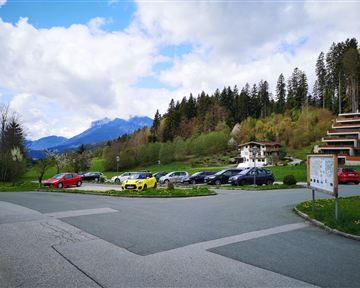 Eine malerische Landschaft mit Bergen im Hintergrund und einem Parkplatz voller Autos. Die Umgebung ist mit Bäumen und Wiesen grün bewachsen.