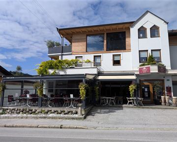 Ein modernes Haus mit einer Terrasse im Erdgeschoss und großen Fenstern. Draußen sind Tische und Stühle für Gäste.