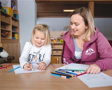 Ein fröhliches Mädchen und eine junge Frau sitzen an einem Tisch und zeichnen. Bunte Stifte und Papiere liegen vor ihnen.