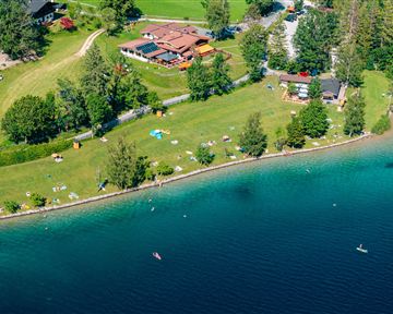 A picturesque lake with green grass and trees along the shore. In the background, there are some buildings and people relaxing by the water.
