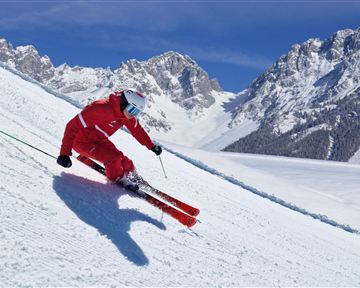A skier in red is skiing down a snow-covered slope. In the background, majestic mountains and a clear blue sky can be seen.