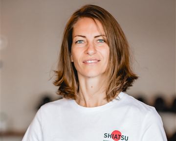 A woman with brown hair is smiling in a bright room. She is wearing a white T-shirt with the print "SHIATSU".