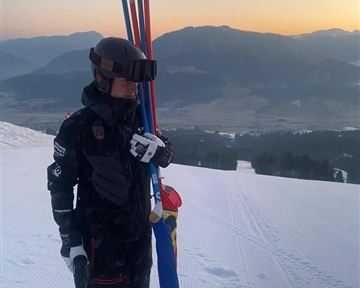 A skier stands on a snowy slope with ski poles in hand. In the background, the mountains and the sunset can be seen.