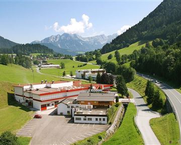 Eine idyllische Landschaft mit grünen Hügeln und Bergen im Hintergrund. Im Vordergrund sind mehrere Gebäude und eine Auto­straße zu sehen.