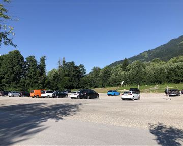 Ein großer Parkplatz, der teilweise gefüllt ist, umgeben von Bäumen und Bergen. Der Himmel ist klar und blau.