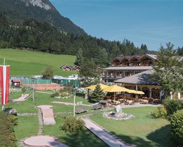 A picturesque hotel complex with a well-maintained garden and a mini-golf course. In the background, green meadows and majestic mountains can be seen.