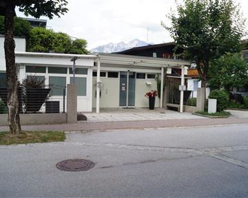 Ein modernes Gebäude mit großen Fenstern und einem Eingangsbereich. Im Vordergrund sind Bäume und eine ruhige Straße sichtbar.