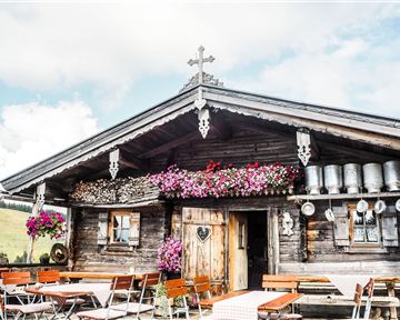 A traditional old wooden house with colorful flowers and friendly outdoor seating. The sky is partly cloudy and the atmosphere is cozy.