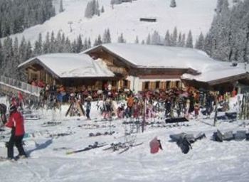 A lively ski hut area in the snow with many skiers and ski equipment. In the background, there are snow-covered trees and mountains.