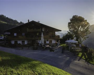 Ein traditionelles Berghaus umgeben von Natur. Die Sonne geht hinter den Bergen auf und beleuchtet die Landschaft.