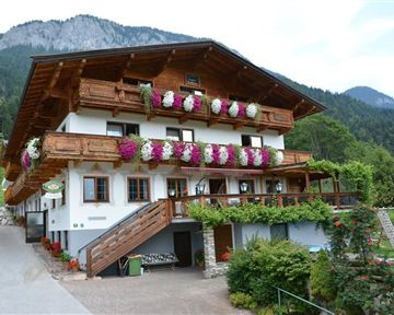 Ein traditionelles alpenländisches Haus mit blühenden Balkonen. Umgeben von grünen Bergen und einer einladenden Gartenlandschaft.