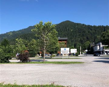 A tranquil landscape with green trees and a view of the mountains. In the foreground, there is a square with unpaved ground and buildings in the background.
