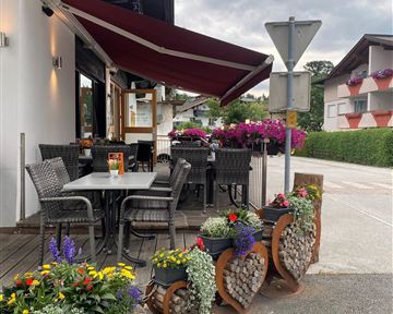 Ein gemütlicher Außenbereich eines Restaurants mit Tischen und Stühlen. Bunte Blumenampeln und dekorative Pflanzen verleihen dem Platz eine einladende Atmosphäre.
