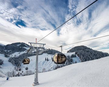 Eine Seilbahn fährt über verschneite Berge. Der Himmel ist teilweise bewölkt und die Landschaft ist winterlich schön.