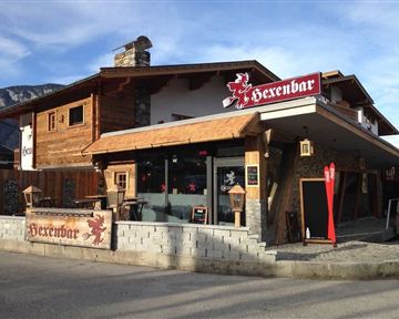 Ein charmantes Holzhaus mit dem Namen "Hexenbar". Es ist eine einladende Location mit einer attraktiven Terrasse und rustikalem Ambiente.