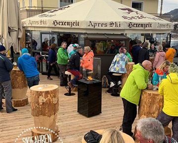 Ein belebter Après-Ski-Bereich mit vielen Menschen, die um Holzstämme und einen Grill versammelt sind. Im Hintergrund ist ein Chalet mit einem großen Sonnenschirm zu sehen.