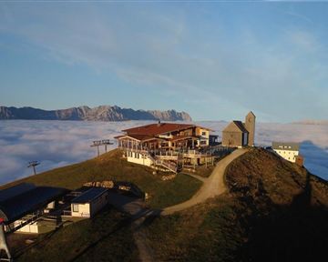 Eine Berghütte liegt hoch über den Wolken mit Blick auf die Berge im Hintergrund. Die Szenerie ist friedlich und bietet eine atemberaubende Aussicht.