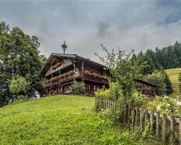 A traditional wooden house surrounded by green meadows and trees. In the background, gentle hills and a cloudy sky can be seen.