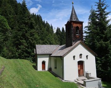 A small church surrounded by dense trees. The sky is blue with some clouds.