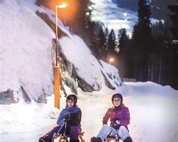 Zwei Personen sitzen auf einem Schlitten in der verschneiten Landschaft. Es ist Abend und die Umgebung ist von Lichtern beleuchtet.