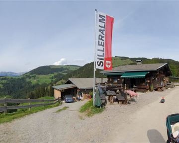 A cozy mountain house with a large flag and a picturesque view of the green hills. The path to the house is lined with trees and invites you to linger.