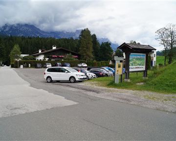 A parking lot in a picturesque setting with mountains in the background. On the left side, there is an information sign visible.