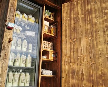 Ein Holzstand mit offener Tür, in dem Milchprodukte und frische Lebensmittel angeboten werden. Der Kühlschrank ist sichtbar und zeigt viele Flaschen in der Auslage.