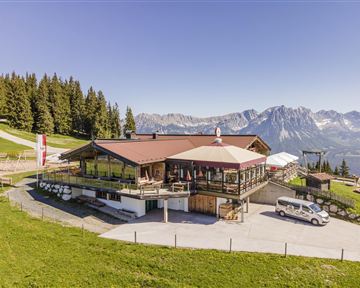 Eine alpine Berghütte mit schöner Aussicht auf die Berge. Umgeben von Bäumen und grünem Gras, bietet sie eine einladende Atmosphäre.