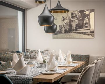 A cozy restaurant with a beautifully set table and elegant chairs. Stylish lamps hang above the table and there is a vintage picture on the wall.