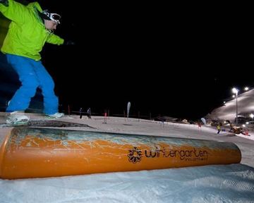 Ein Snowboarder springt über ein oranges Rail bei Nacht. Die Piste ist beleuchtet und bietet eine winterliche Atmosphäre.