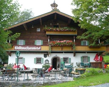 A traditional wooden house with blooming balconies and an inviting terrace. Guests sit outside and enjoy the peaceful surroundings.