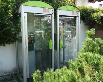 Two phone booths stand next to each other, surrounded by green plants. The phone booths have a modern shape and are partially equipped with glass fronts.