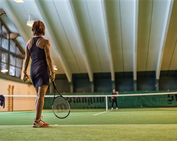 A female tennis player stands on the court and looks towards the net. The interior is well lit with a green playing surface.
