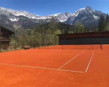A tennis court with a red surface, surrounded by majestic mountains and a clear blue sky. Nature adds a picturesque backdrop.