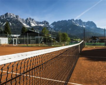 A tennis court with red clay and a clear blue sky. Impressive mountains rise in the background.