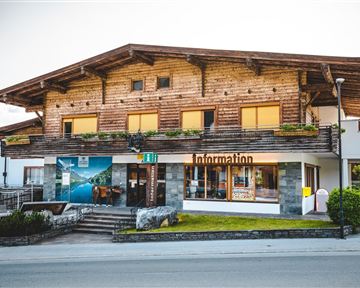 Ein traditionelles Holzgebäude mit einem Schild "Information" an der Vorderseite. Es liegt an einer Straße und hat große Fenster, die von Blumen umgeben sind.