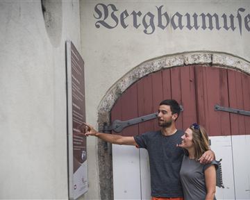 A couple stands in front of the entrance of the mining museum and looks curiously at a sign. The atmosphere is relaxed and inquisitive.