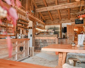Ein gemütlicher Raum mit großen Fenstern, der einen Blick auf verschneite Häuser und Bäume bietet. Am Tisch stehen Holzstühle und eine dekorierte Tischdecke, während Regale mit verschiedenen Produkten gefüllt sind.