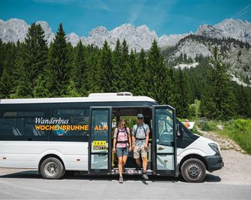 Ein Wanderbus steht in der Natur, umgeben von Bergen und Wäldern. Zwei Personen steigen aus, bereit für ein Abenteuer.