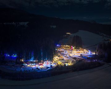 Eine festlich beleuchtete Winterlandschaft mit einem kleinen Dorf inmitten von schneebedeckten Bäumen. Die Lichter schaffen eine gemütliche Atmosphäre in der Nacht.