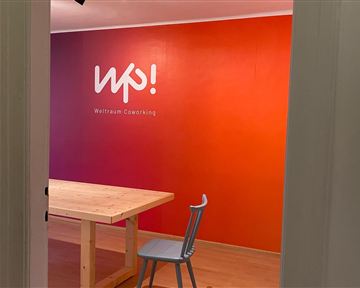 Ein freundlicher Coworking-Raum mit einem großen Tisch und einem Stuhl. Die Wand ist in warmen Farben gestaltet und trägt das Logo „wp!“.