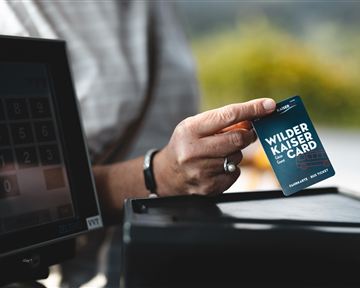 A person is holding a Wilder Kaiser Card in their hand. In the background, a cash register terminal is visible.