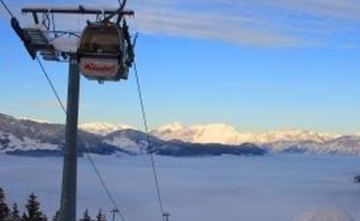 Eine Seilbahn schwebt über eine bewaldete Berglandschaft. Im Hintergrund sind schneebedeckte Berge und ein Nebelmeer zu sehen.