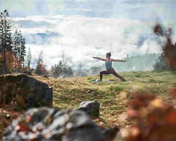 A person is doing yoga in a picturesque, wooded landscape. Gentle hills and mist can be seen in the background.