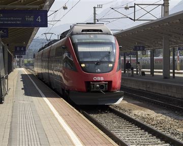A red train of the ÖBB is at the station. The tracks are visible and the architecture of the station is modern.