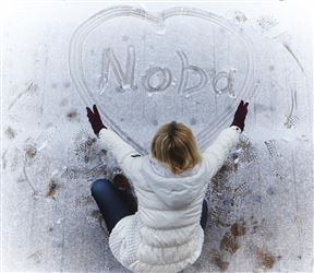 Eine Person sitzt auf dem Schnee und schreibt den Namen "Noba" in ein Herz. Sie trägt eine hellgraue Jacke und rote Handschuhe.