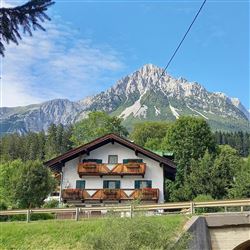 A cozy house with balconies, surrounded by lush greenery. In the background, an impressive mountain range rises under a clear blue sky.