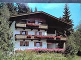 A traditional chalet with blooming balcony boxes and a well-maintained garden. Surrounded by trees and a clear blue sky.