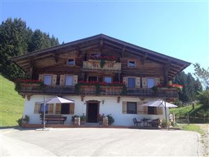 A traditional chalet with a wooden facade and flowers on the balcony. In the foreground, there are tables and sun umbrellas.